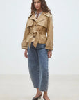 A woman with long wavy hair and a neutral expression stands in a minimalist room wearing the SUNCOO Elliott Jacket over a brown polka dot top, blue high-waisted jeans, and black pointed-toe heels.