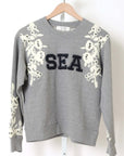 The Georgie Sweatshirt by Sea New York is a gray cotton-polyester blend featuring bold black "SEA" lettering, white floral appliqués, and black beads on the shoulders and sleeves. Displayed on a wooden hanger. True to size; dry clean recommended.