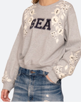 A person wears the Sea New York Georgie Sweatshirt—a light gray, true to size cotton blend with "SEA" in bold black letters, white floral embroidery and sequins—paired with distressed blue denim jeans.