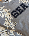The Georgie Sweatshirt by Sea New York is a gray cotton-poly blend featuring "SEA" in large navy letters, white embroidered flowers, and black bead details on the sleeve and shoulder. True to size—perfect for any casual look.