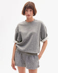 A woman with short brown hair wears the Clare Vivier SS Oversized Sweatshirt in gray cotton fleece with rolled sleeves, paired with matching shorts, standing against a white background and smiling softly.