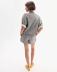 A person with short brown hair and yellow flats stands against a white background, wearing the Clare Vivier SS Oversized Sweatshirt— a loose-fitting, gray speckled 100% cotton top with rolled sleeves, paired with matching shorts.
