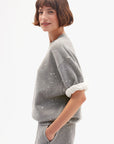 A person with short brown hair wears the Clare Vivier SS Oversized Sweatshirt, a grey soft cotton fleece top with white speckles, styled with matching pants. They stand sideways with one hand in their pocket and a rolled-up sleeve.