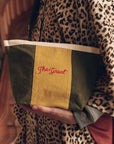 The Striped Pouch in olive green and yellow cotton canvas with a cream band and red logo held in hands.