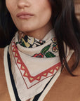 A woman wears The Derby Silk Bandana by The Great Inc., featuring a vintage light floral design around her neck, paired with a tan sweater with black and white trim. Only the lower half of her face and neck are visible.