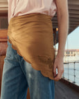 A person stands on a wooden deck near the water, wearing blue jeans, a light pink shirt, and The Scalloped Bandana skirt with vintage handkerchief-inspired floral embroidery by The Great Inc.