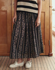 A person stands on a wooden floor wearing a black and cream striped sleeveless top, white boat shoes, and The Canary Skirt by The Great Inc., an airy pull-on skirt with floral and stripe patterns. The person's face is not visible.