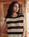 A woman with long dark hair stands indoors in front of a wooden door, wearing The Great Inc.'s Crochet Sweater Shell—a sleeveless black and beige striped crochet top with vintage sailor-inspired style. She gazes thoughtfully to the side.