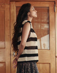A woman with long dark hair stands in profile before wooden doors, wearing The Crochet Sweater Shell by The Great Inc., her hands clasped behind her back.