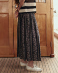 A person stands indoors on a wooden floor, wearing The Canary Skirt by The Great Inc.—an airy pull-on skirt with floral designs and vertical stripes—paired with a black and white striped sleeveless top and white boat shoes. Their face is out of the frame.