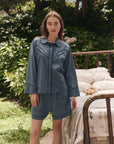 A woman poses outdoors beside a brass bed, wearing The Great Inc.'s The Pajama Shirt and matching shorts. Surrounded by greenery and flowers, she exudes relaxed nostalgia in this natural setting.