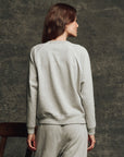 A person with long brown hair wears The Fleece Rookie Sweatshirt by The Great Inc., made in Los Angeles, and matching sweatpants, standing with their back to the camera against a textured dark background.