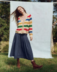 On sunny grass, a person wears a colorful striped cardigan, The Rhythm Skirt by The Great Inc. in long blue cotton eyelet style, and brown boots. They're framed by trees with a light blue backdrop, exuding Victorian charm.