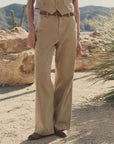 In a desert landscape among rocks and plants, a person wears The Mill Pant by The Great Inc.—tan cotton herringbone carpenter pants—paired with a matching vest, brown belt, and brown shoes, with their upper body partially out of frame.