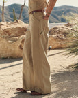 A person stands outdoors on sandy ground wearing The Great Inc.'s The Mill Pant—wide-legged, high-waisted cotton herringbone workwear pants—with brown shoes, one hand in their pocket and rocky hills in the background.