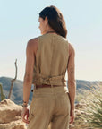 A woman stands outdoors with her back to the camera, wearing The Suit Vest and matching pants by The Great Inc. in light brown cotton herringbone, set against a desert landscape with cacti and rocky hills.