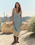 A woman stands in a desert landscape wearing The Legacy Dress by The Great Inc.—a 1920s inspired light blue silk dress with cream embroidered mesh lace trim—paired with brown cowboy boots, surrounded by rocks, cacti, and mountains.
