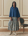 A woman stands indoors beside a vintage chair, wearing The Highland Skirt by The Great Inc.—a floral pleated skirt with an elastic waistband—paired with a loose blue top and light lace-up boots, against cream paneled walls and a patterned rug.