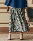 Indoors, a person wears a blue top with The Highland Skirt by The Great Inc.—a blue floral pleated piece featuring yellow and white flowers—paired with beige socks, white ankle boots, and a vintage-style chair nearby.