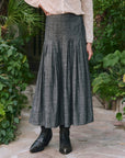 A person wearing The Great Inc.'s The Rhythm Skirt, a high-waisted gray midi, paired with a cream lace blouse and black boots, stands on a stone path among green plants.