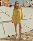 A woman stands on a wooden dock in front of a white boat, wearing The Micro Terry Swing Short by The Great Inc. with a loose-fitting yellow cotton micro terry long-sleeve lace-up top and beige boat shoes.