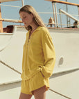 Wearing The Great Inc.'s The Micro Terry Swing Short, a woman in yellow stands before a white boat on a sunny day, hands in her pockets, looking down.