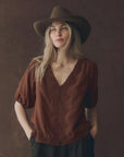 A woman with long blonde hair wears The SS Legacy Top by The Great Inc., a brown silk V-neck top with embroidered mesh lace, paired with a brown wide-brimmed hat and standing against a dark textured background.