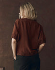 A person with blonde hair, seen from behind, wears The SS Legacy Top by The Great Inc.—a short-sleeved, dark red silk blouse with embroidered mesh lace trim—paired with black pinstripe pants against a 1920s loungewear-inspired backdrop.