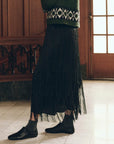 The Fringe Skirt in black tiered fringe styled with a green patterned sweater, dark socks, and black shoes.