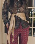 Beige The Wrap Scarf tied at the waist over a patterned button-down and red geometric trousers.