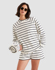 A woman wearing the Kule Terry Franny sweatshirt with matching striped shorts and sunglasses holds a rolled-up striped towel against a plain light gray background.