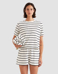 A woman wears Kule’s The Terry Hurley—matching white cotton-blend cropped tee and shorts with black stripes—while holding a rolled-up striped towel against a plain light gray background.