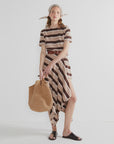 A woman stands against a white background wearing The Terry Hurley dress by Kule, a brown belt, headscarf, black sandals, and holding a large woven tote bag.