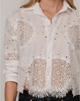 A woman wears The Belle shirt by Cissa—a white, long-sleeve, button-up Egyptian cotton top with intricate lace cut-out details and a scalloped hem—paired with dark denim jeans for an effortlessly elegant look.