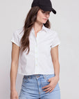 A person wears a black baseball cap, light blue jeans, and THE SHIRT’s The Boxy Short Sleeve Cropped Shirt with No Gape button technology, standing against a plain light background and looking down with one hand in their pocket.