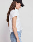 A person with long brown hair wears THE SHIRT's The Boxy Short Sleeve Cropped Shirt, paired with a black baseball cap and light blue jeans, standing in side profile against a plain light background.
