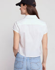 A woman with long brown hair is seen from the back wearing a black baseball cap and THE SHIRT’s Boxy Short Sleeve Cropped Shirt, featuring No Gape button technology, paired with light blue jeans against a plain light background.