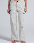 A person stands barefoot, shown from the waist down against a plain light background, wearing ASKK’s Tomboy Twill high-waist straight-leg pants in light beige with blue paint splatters, crafted from stretch twill.
