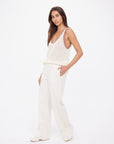 A woman with long brown hair stands against a white background wearing The Upside’s NAVAR MARNIE WIDE LEG PANT in cream and a loose, sleeveless cream knitted jacquard top; she has her hands in her pockets and a neutral expression.