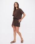 A woman with long brown hair models The Upside's SOHO DOLCI SHORT—a loose, high-rise, wide leg brown short—paired with a matching T-shirt and black flip-flops, posing against a plain white background.