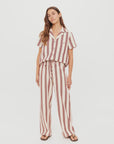 A woman stands against a plain white background wearing The Upside's cream short-sleeve button-up shirt and BAJA CHARLI DRAWSTRING PANT in burnt red stripes, paired with sandals.