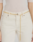 A person wears high-waisted, off-white pants with The Sebastian Belt Gold by FRAME looped through the belt loops and hanging down one side with a tassel detail, paired with a light beige ribbed top.