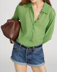 A woman wears The Collared Cardi by FRAME—a sheer green button-up styled like a lightweight knit—paired with denim shorts, a black belt, and carrying a brown leather clutch under her arm. Her head is slightly tilted, face partly visible.