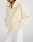 A person wears the FRAME Seersucker Track Jacket in light yellow, featuring snap buttons and a high collar, paired with white pants. Their hands are in their pockets, and their face is partially out of frame.