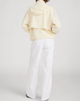 A person stands with their back to the camera, wearing FRAME's The Seersucker Track Jacket in light yellow with a vented back and loose white pants. The lightweight jacket pops against the plain white background.