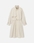 The Field Trench by FRAME is a cream-colored cotton coat featuring a belted waist, asymmetric wide lapel, buttoned cuffs, and two side pockets, shown against a plain white background.