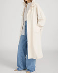 A woman wears The Field Trench by FRAME—a long, loose, cream cotton coat—over a light shirt and wide-leg blue jeans with beige pointed-toe shoes. One hand is in her pocket and her head is turned slightly to the side.