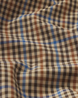 A close-up of The Plaid Mac Coat by FRAME shows its structured cotton-blend fabric in black, brown, blue, and beige checks with gentle folds and shadows on the slightly wrinkled surface.