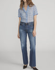 A person stands against a plain background wearing the FRAME Essential Top in blue plaid, paired with blue high-waisted jeans and black pointed-toe shoes. Their wavy hair falls loosely around their shoulders.