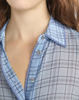 A close-up of a person in the FRAME Essential Top, a lightweight, button-front plaid shirt in light blue with gray and navy lines. The shirt is partially unbuttoned, showing wavy brown hair.